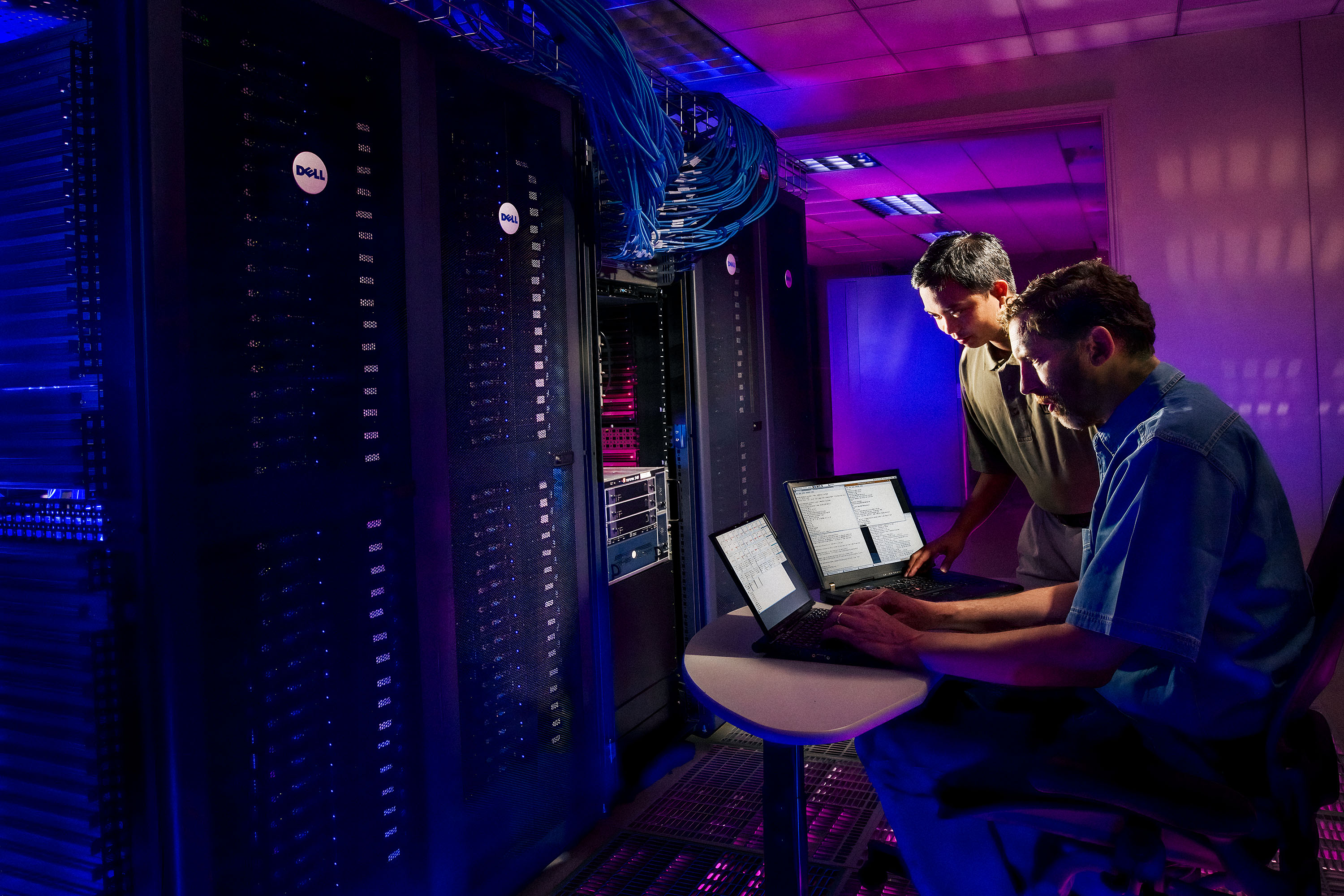 Two men working on laptops in a data center setting. The light is blue and purple.  