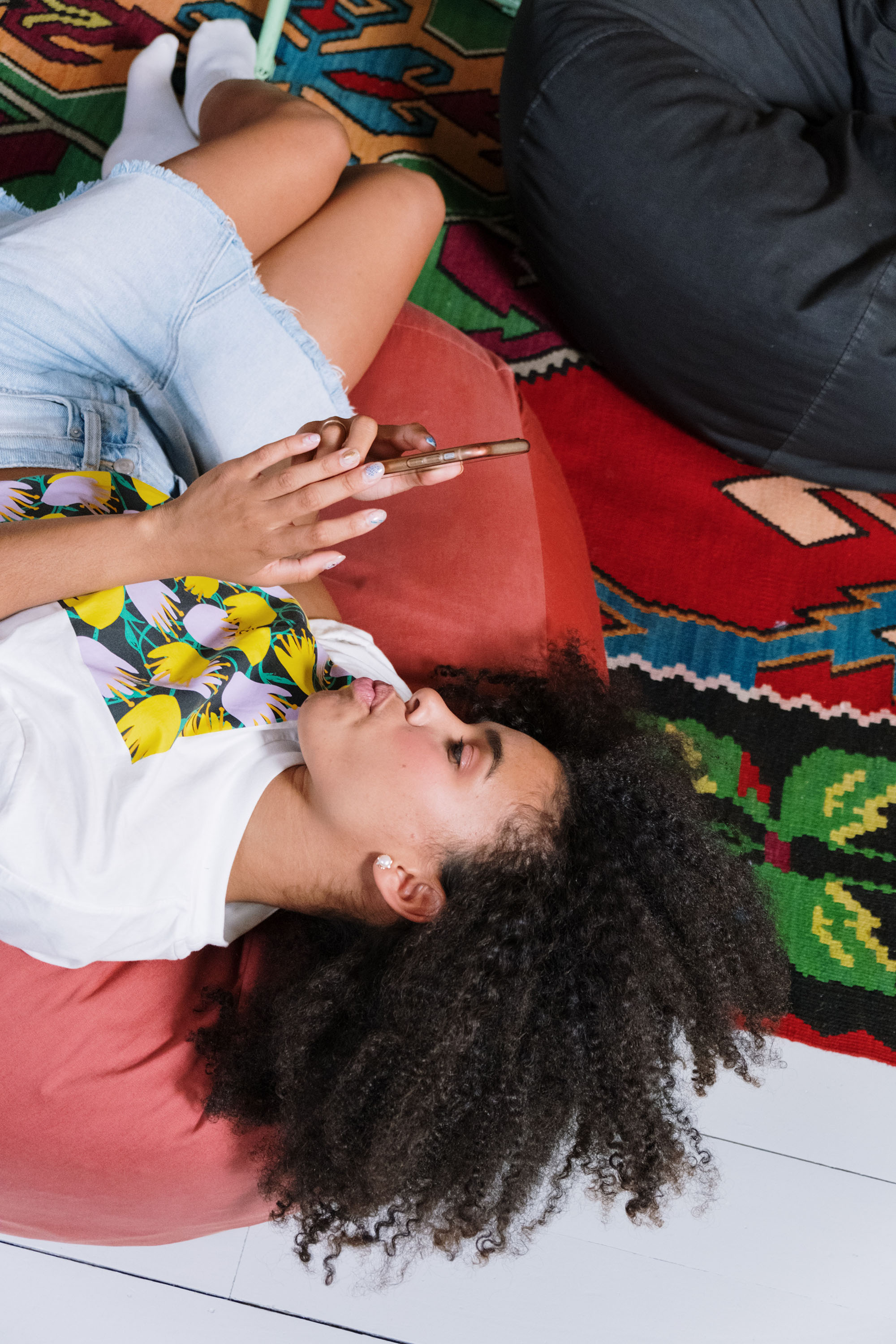 A person is lying on a vibrant rug, head on a red cushion, wearing a floral shirt and denim shorts, holding a smartphone.
