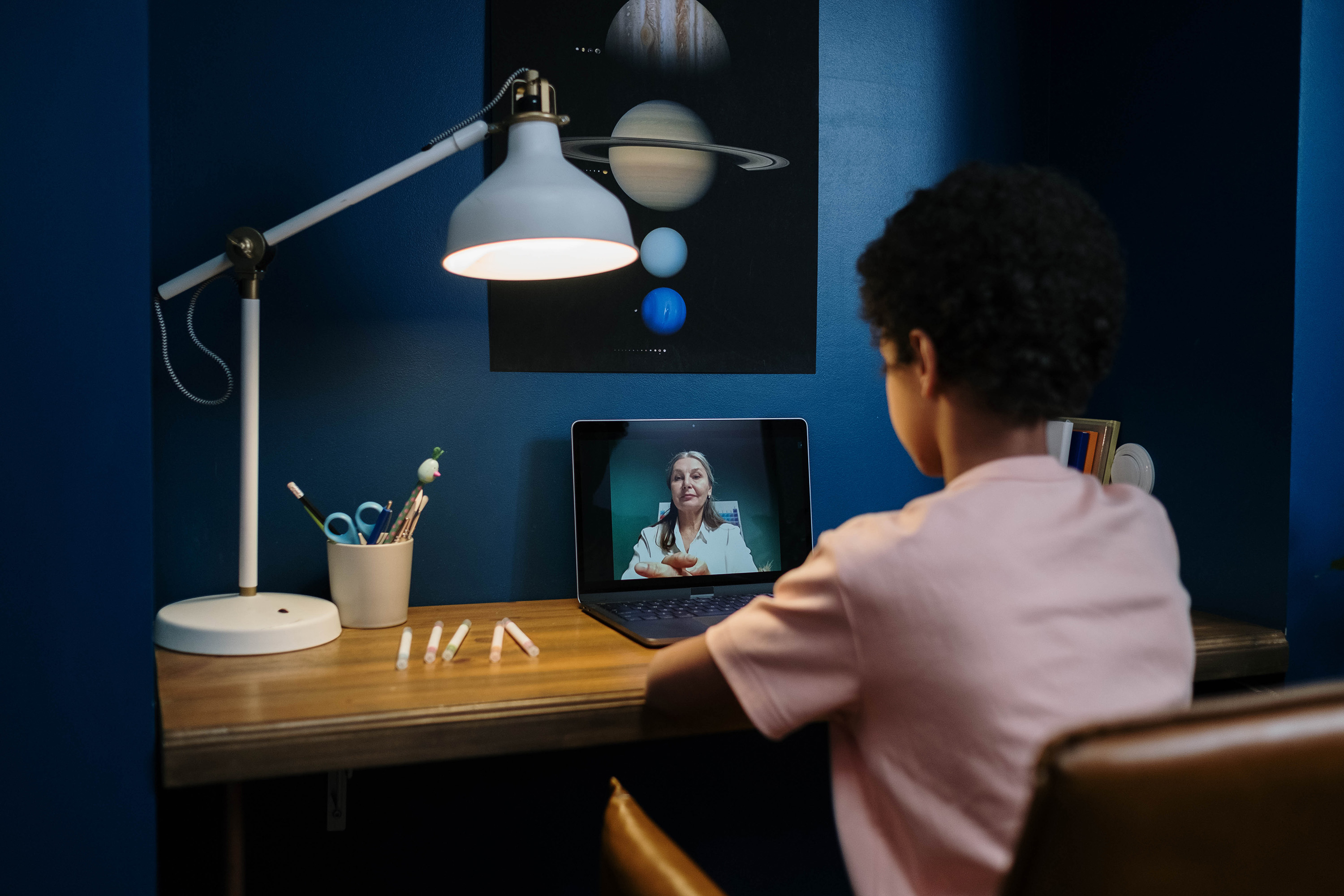 A person is at a desk under a lamp, in a virtual meeting on a laptop, surrounded by stationery and a poster of planets.